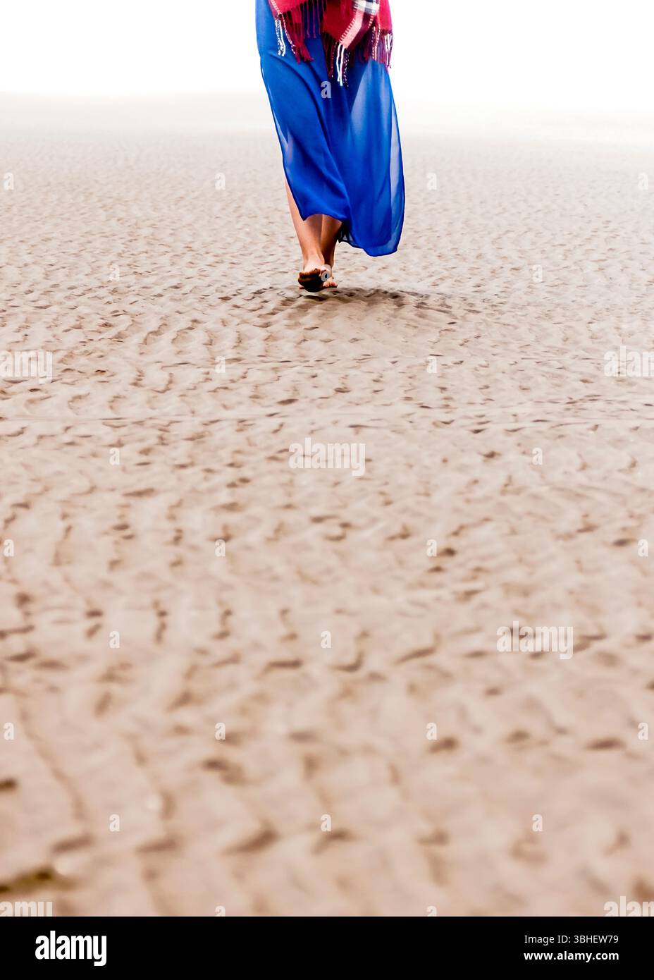 Une femme portant une robe bleue et un châle à carreaux rouge et blanc marche pieds nus sur une plage de sable. Banque D'Images