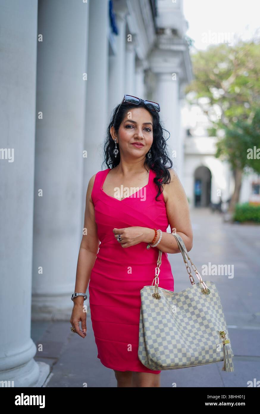 Femme élégante dans une robe rose vif portant un sac à main designer tout en marchant à travers une promenade de la ville, mode et concept de style de vie urbain moderne. Banque D'Images
