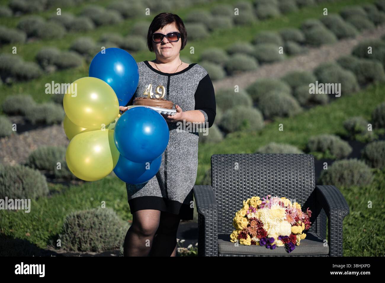 Une femme tient joyeusement un gâteau orné d'un numéro et de ballons dans un beau champ de lavande, célébrant son anniversaire dans un cadre extérieur serein f Banque D'Images