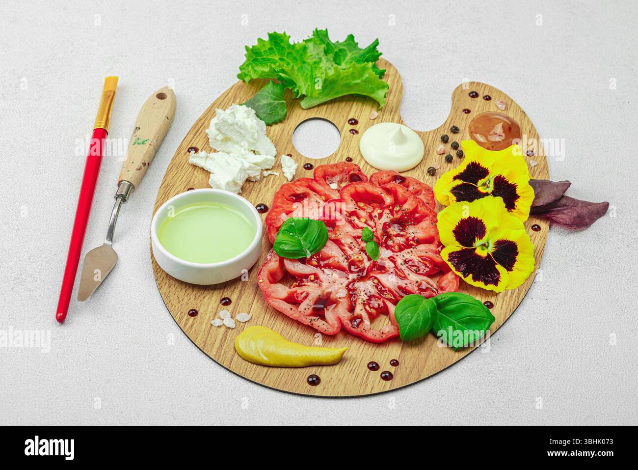 Tomate héritée tranchée avec feta, herbes, fleurs comestibles et sauces sur une palette d'artistes en bois, stylisée avec pinceau et couteau à palette. Concr. Pierre claire Banque D'Images