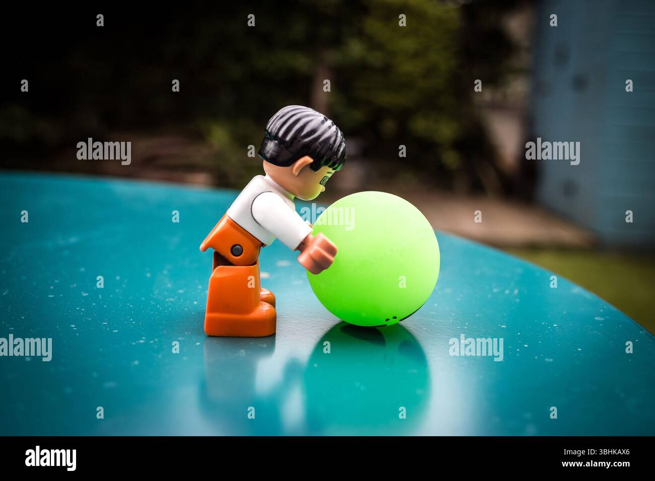 Jouet Lego figure d'une personne en tenue orange et blanche jouant avec une balle verte sur une surface brillante à l'extérieur - concept de jeu de temps de jeu Banque D'Images