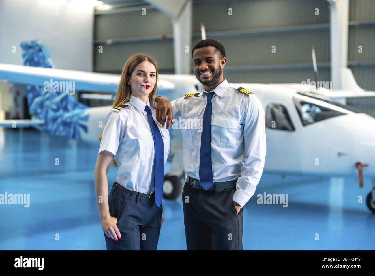 Deux pilotes de ligne souriants posant en toute confiance devant un jet privé à l'intérieur d'un hangar spacieux, mettant en valeur leur camaraderie professionnelle Banque D'Images