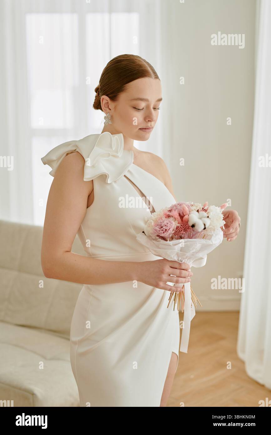 Femme jeune adulte caucasienne debout avec les yeux fermés tenant le bouquet de mariage dans les mains portant une robe élégante dans une pièce lumineuse pendant la célébration de mariage Banque D'Images