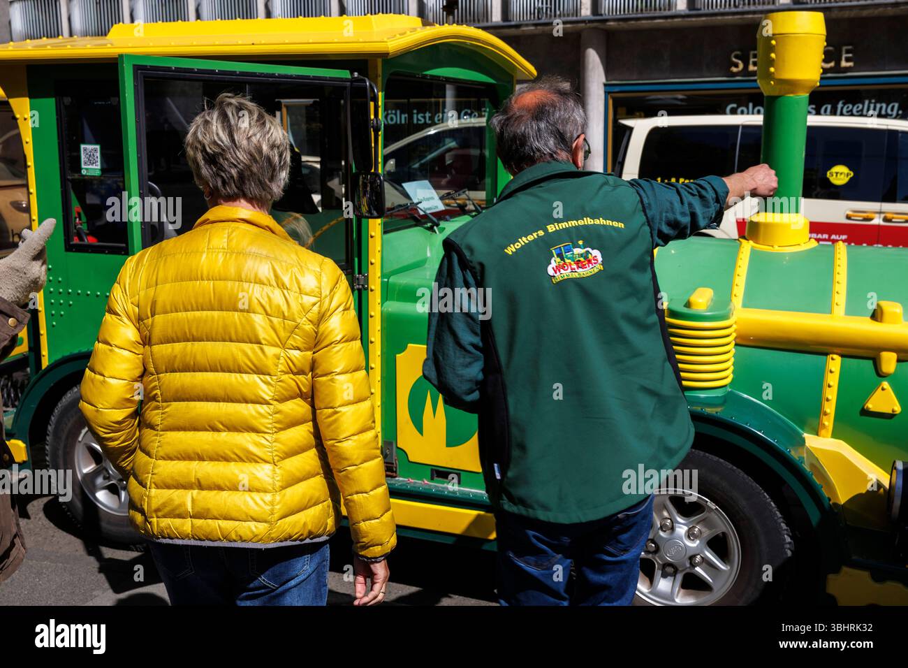 Femme en veste jaune se tient devant le mini train touristique de Cologne et a quelque chose expliqué par le chauffeur, Cologne, Allemagne. Frau in gelb Banque D'Images