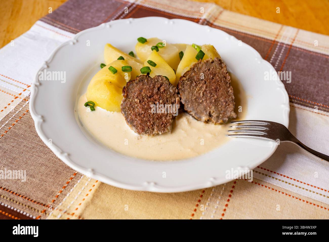 Deux morceaux de pain de viande cuit au four, pomme de terre avec ciboulette sur assiette blanche, sauce raifort, fourchette sur nappe à carreaux. Banque D'Images