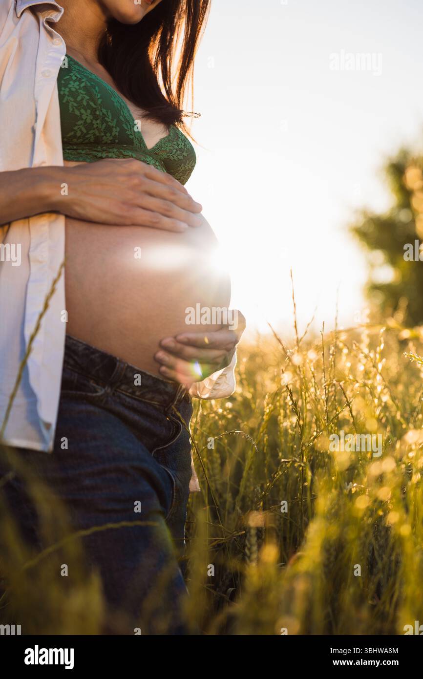 Gros plan d'une future mère en soutien-gorge en dentelle verte et jeans debout dans un champ doré, berçant doucement son ventre avec un contre-jour chaud du cadre Banque D'Images