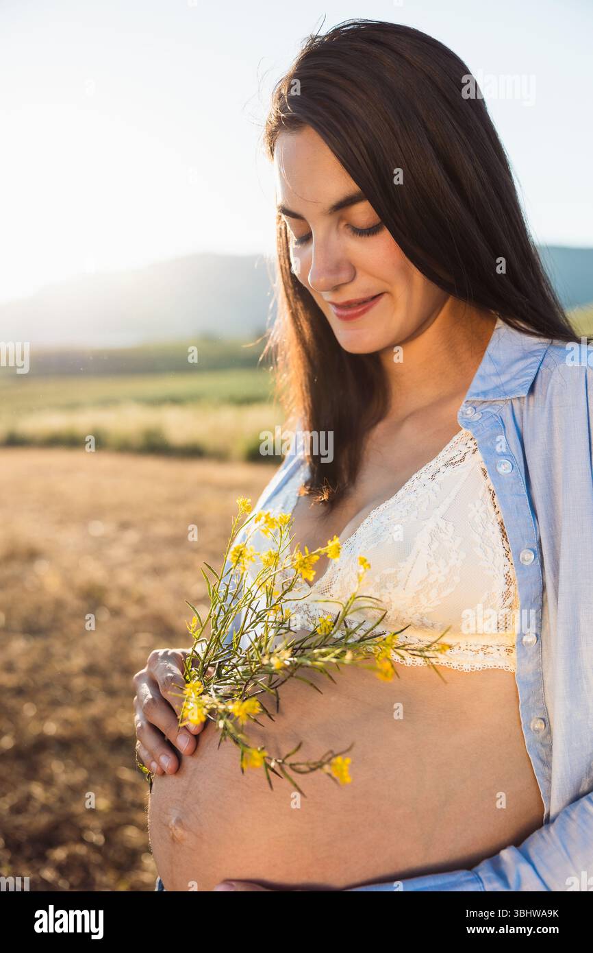 Future mère dans un soutien-gorge en dentelle blanche et chemise bleue sourit doucement tout en tenant un bouquet de fleurs sauvages jaunes sur son ventre enceinte dans un co doré Banque D'Images