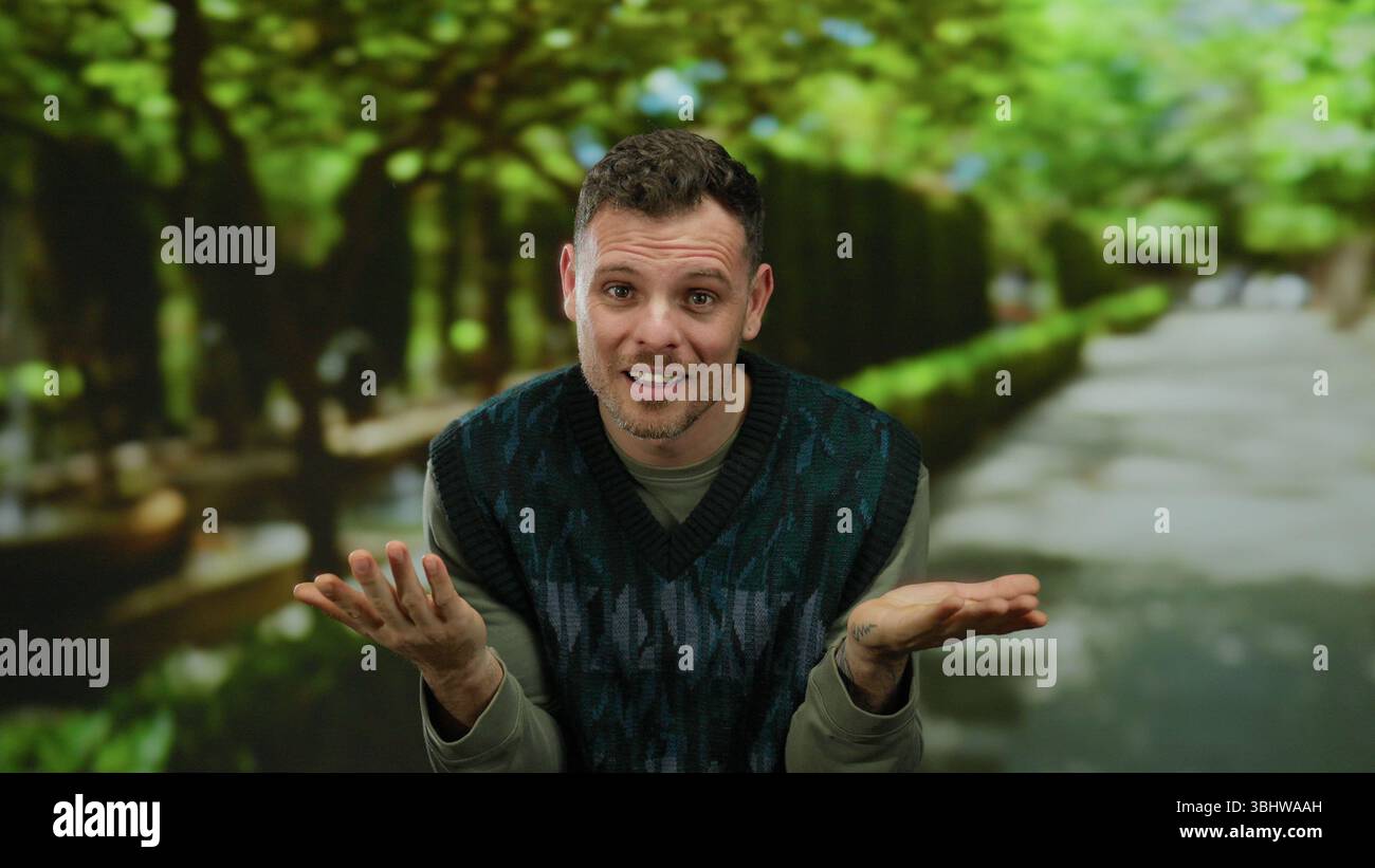 Homme haussant les épaules à l'extérieur vert parc tenue décontractée lumière du jour arbres flou arrière-plan détendu ambiance réfléchie portrait franc geste émotion naturel envi Banque D'Images