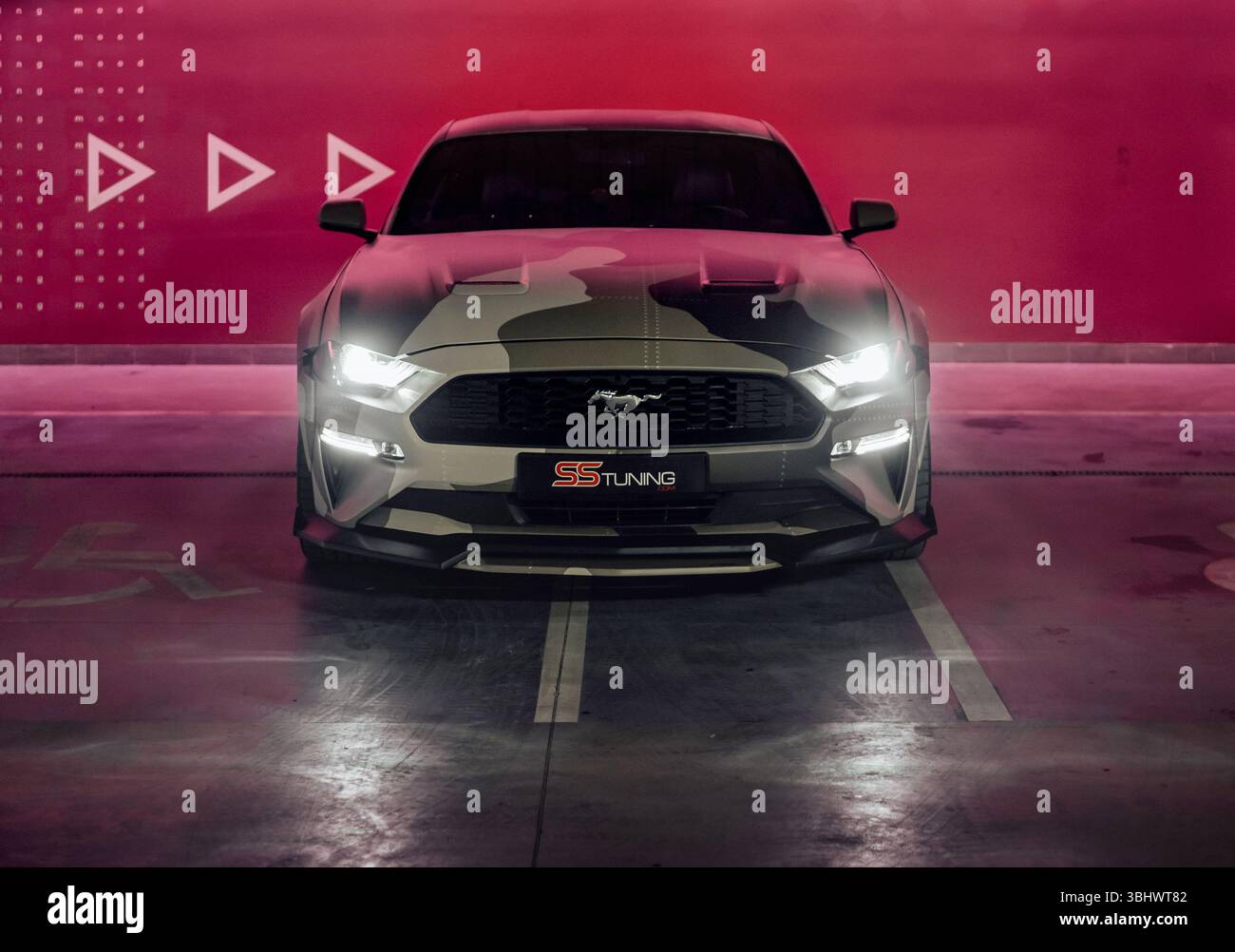 Camo beige Ford Mustang dans le garage de stationnement. Vue avant du poney car américain garé près du mur rouge illuminé. Banque D'Images
