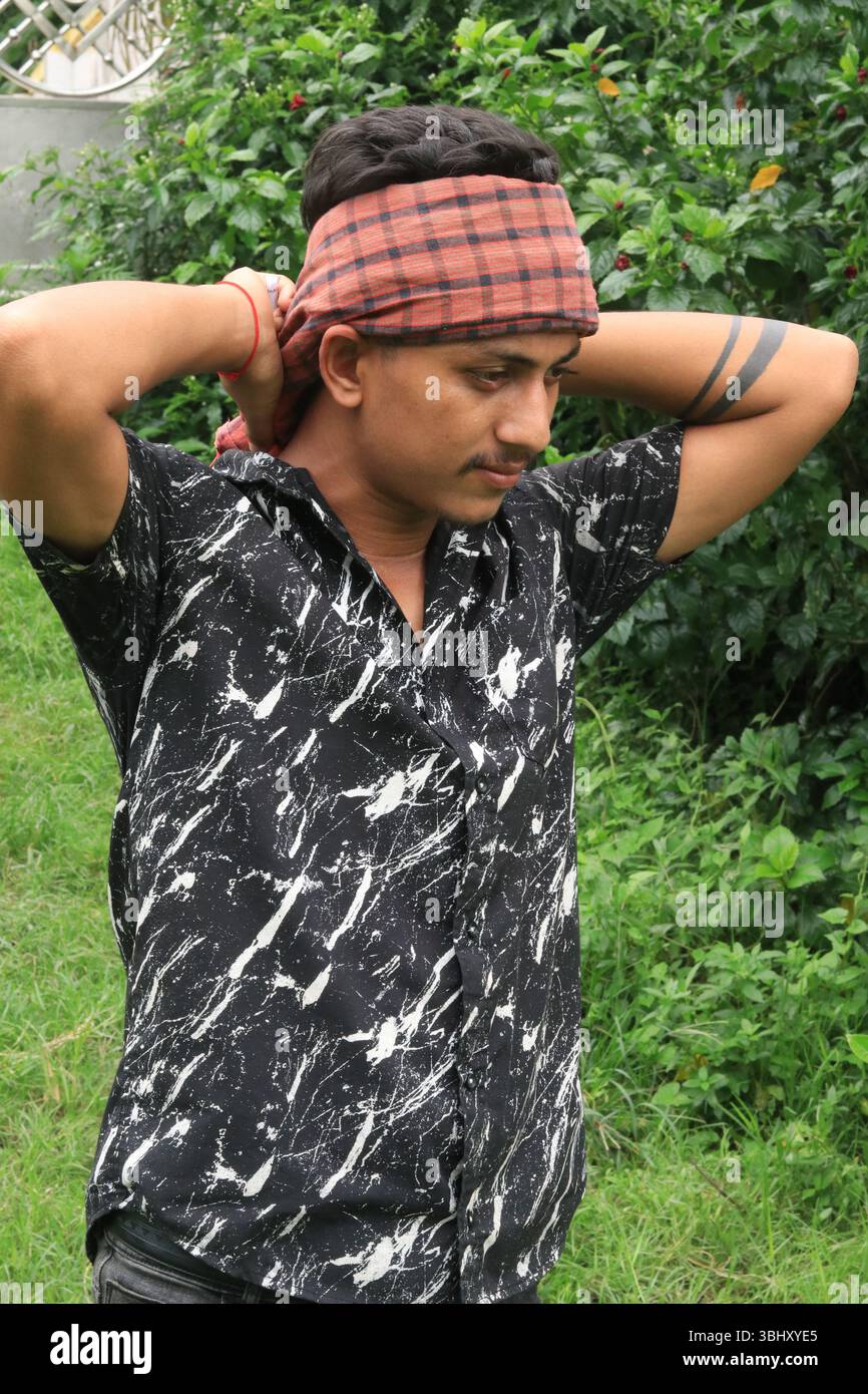Jeune ouvrier sud-asiatique avec un bandana rouge traditionnel (gamcha) attaché sur le front, posant la main sur la tête. Concept de travail acharné et rural l Banque D'Images