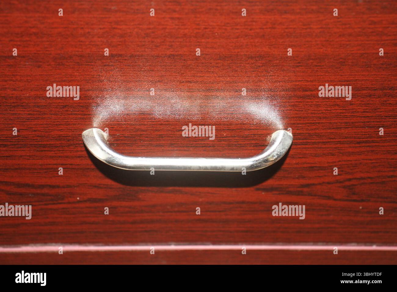 Vue rapprochée d'une poignée de tiroir métallique montée sur une surface en bois poli d'une façade de tiroir. Le bois a une teinte rougeâtre-brun riche. Banque D'Images