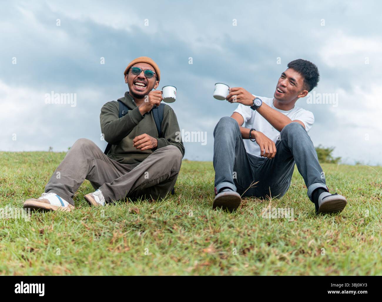 Deux hommes joyeux assis sur une colline herbeuse, riant et riant des tasses en émail. Un joyeux moment d'amitié, de voyage et de loisirs en plein air capturé dans la nature. Banque D'Images