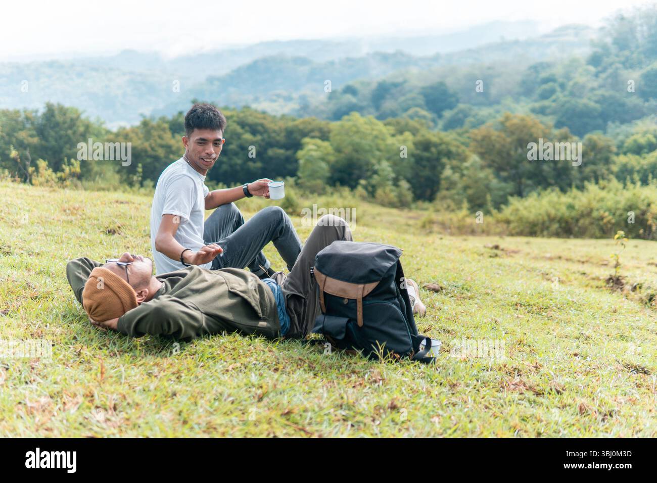 Deux hommes indonésiens se détendent sur une colline herbeuse, riant et dégustant une tasse de café avec une forêt pittoresque et une montagne en toile de fond. Banque D'Images