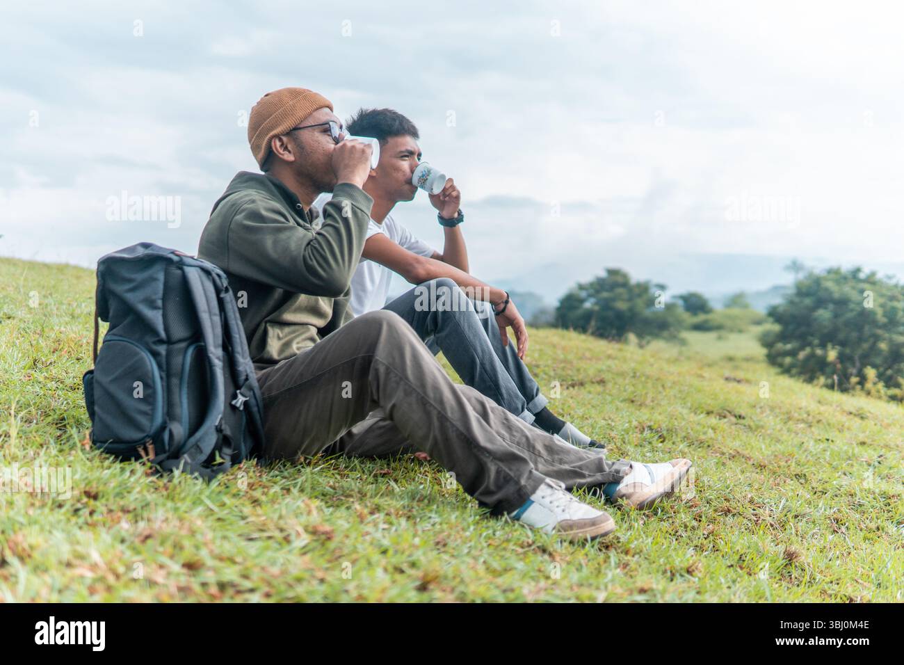 Deux hommes joyeux assis sur une colline herbeuse, riant et riant des tasses en émail. Un joyeux moment d'amitié, de voyage et de loisirs en plein air capturé dans la nature. Banque D'Images