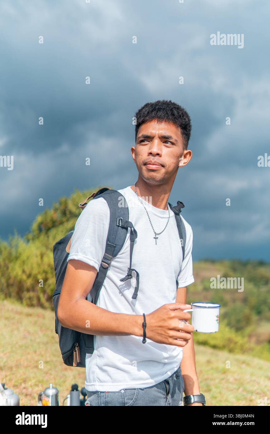 Un homme indonésien avec un sac à dos et un bonnet déguste une tasse de café tout en regardant au loin. Banque D'Images