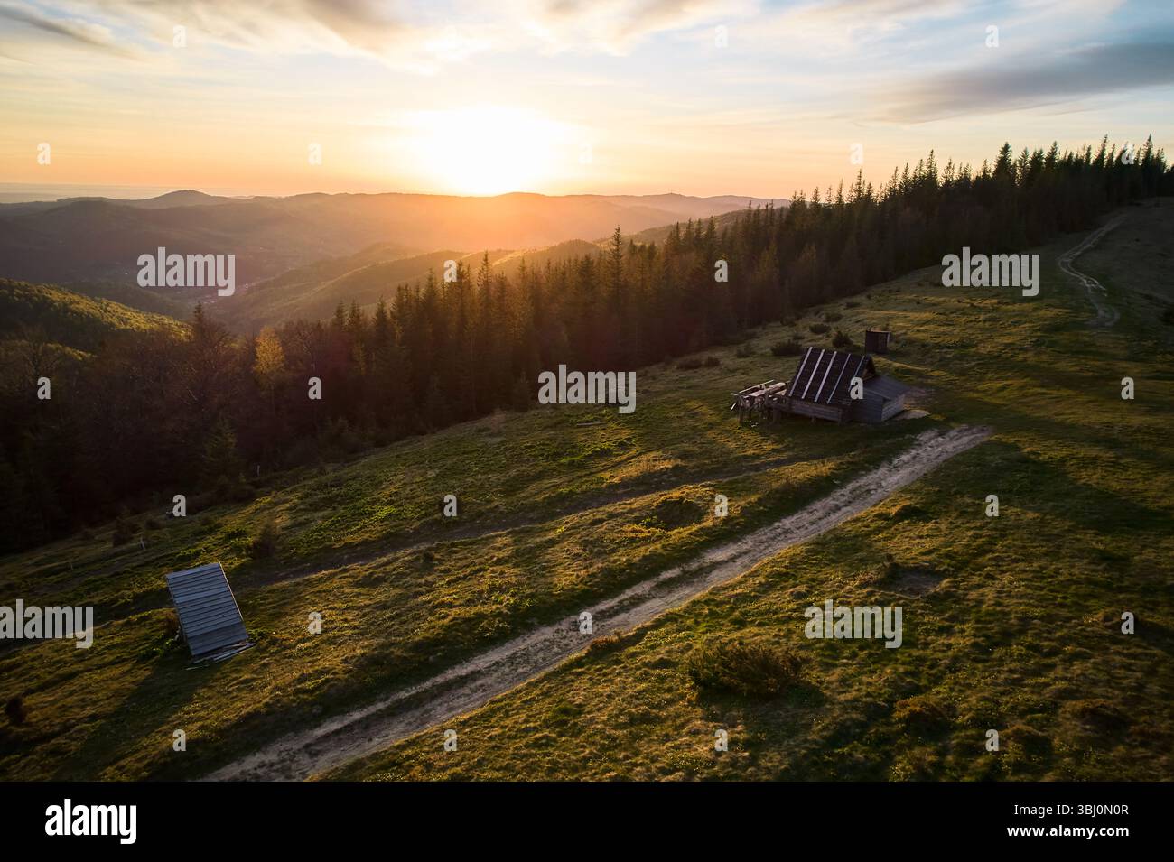 Vue aérienne du coucher de soleil serein sur un paysage vallonné en Ukraine. Cabane en bois rustique et chemin de terre niché parmi les collines verdoyantes, avec la forêt et les montagnes baignées de lumière chaude et dorée. Banque D'Images