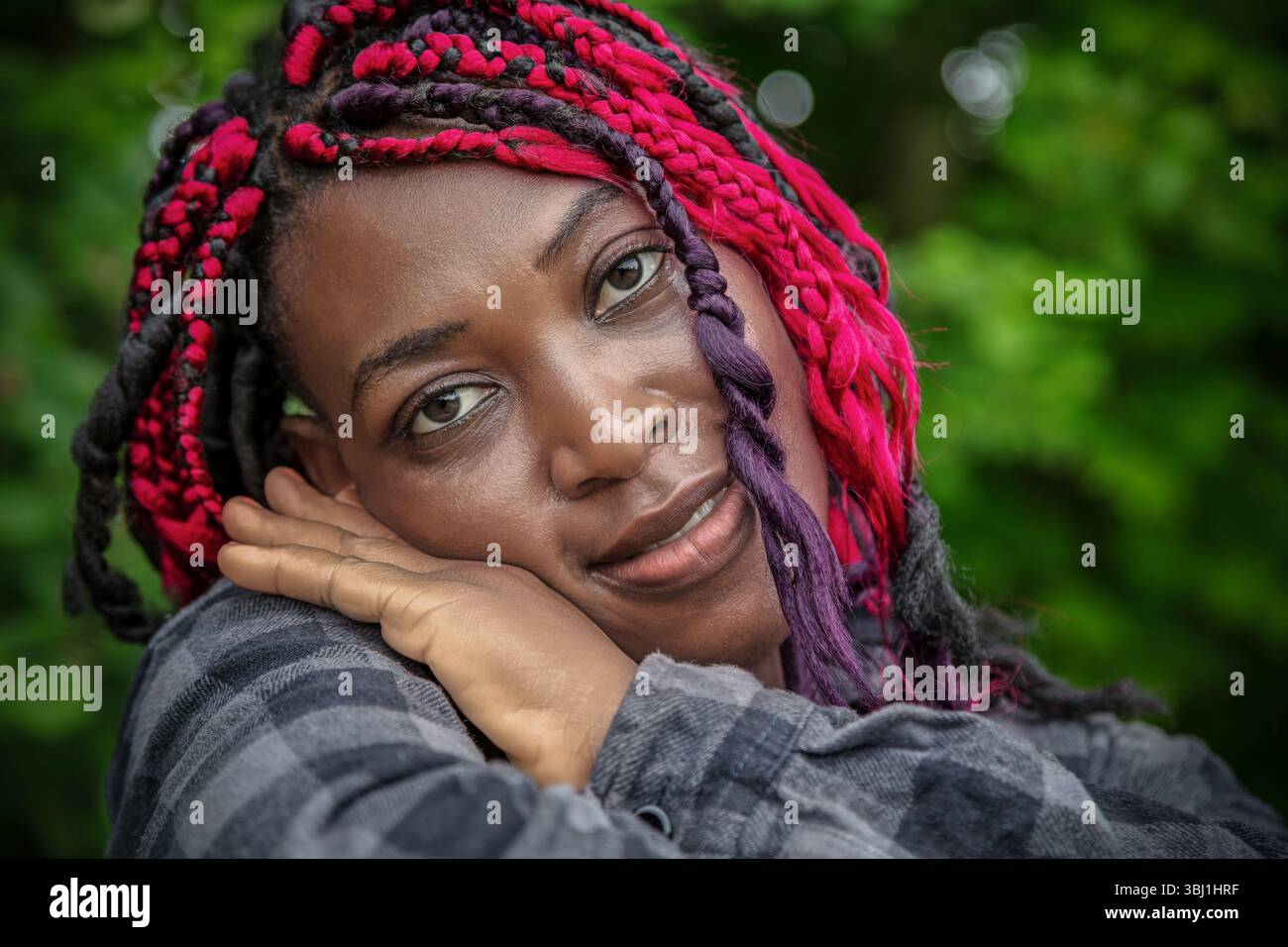 Charmante femme afro-américaine avec coiffure tressée rose reposant la tête sur les mains dans le parc vert. Portrait naturel du style de vie en extérieur avec un sourire authentique Banque D'Images