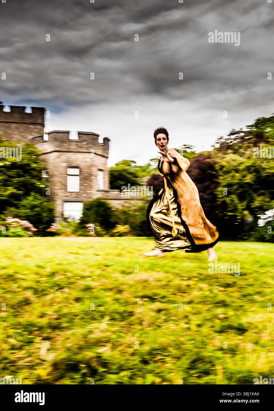 Une femme vêtue d'une robe d'or court à travers un champ herbeux devant un château. Banque D'Images