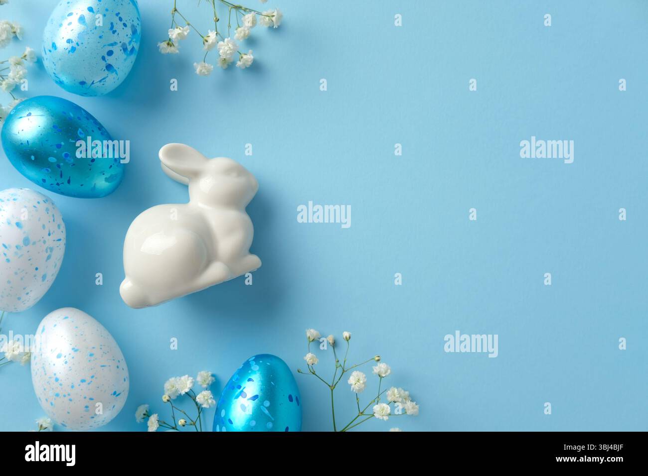 Lapin en céramique blanc parmi les œufs de Pâques mouchetés bleus et blancs et délicates fleurs blanches sur un fond bleu clair Banque D'Images