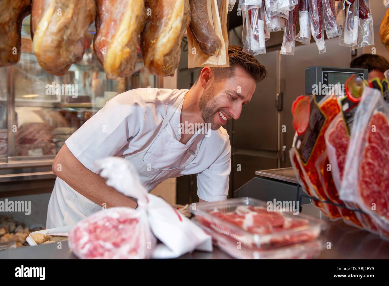 Boucher souriant tout en préparant habilement des filets de viande fraîche dans une boucherie animée, mettant en valeur les compétences et le dévouement à la qualité Banque D'Images
