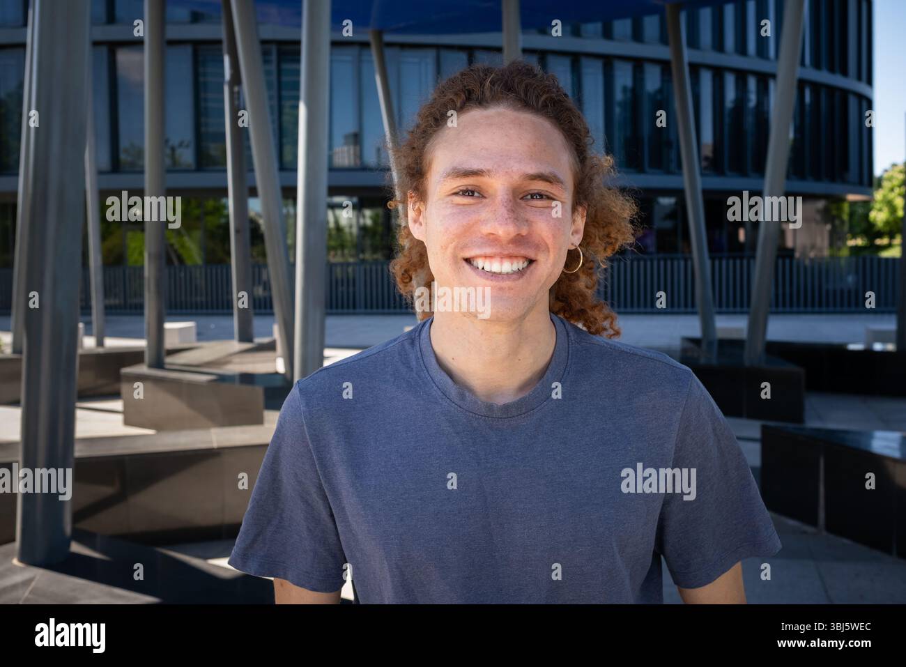 Jeune homme souriant avec des cheveux bouclés et boucle d'oreille debout à l'extérieur dans un environnement urbain moderne. Style décontracté, journée ensoleillée, concept de style de vie contemporain. Banque D'Images