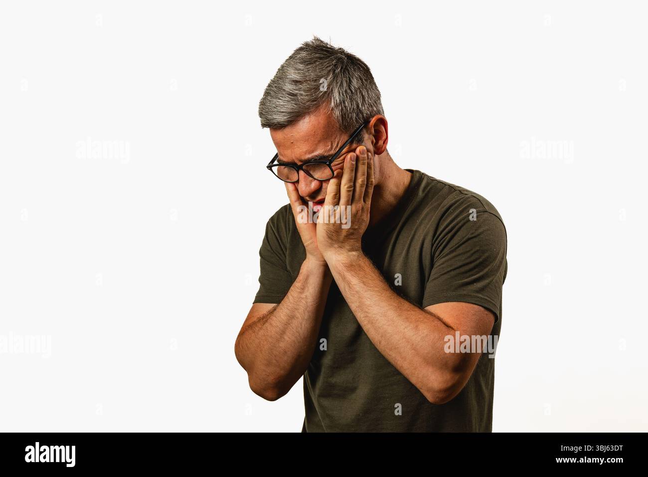 Un homme debout, les mains sur son visage, véhiculant des émotions de détresse ou de contemplation dans un cadre intérieur légèrement éclairé. Ses cheveux gris ajoutent à la t Banque D'Images