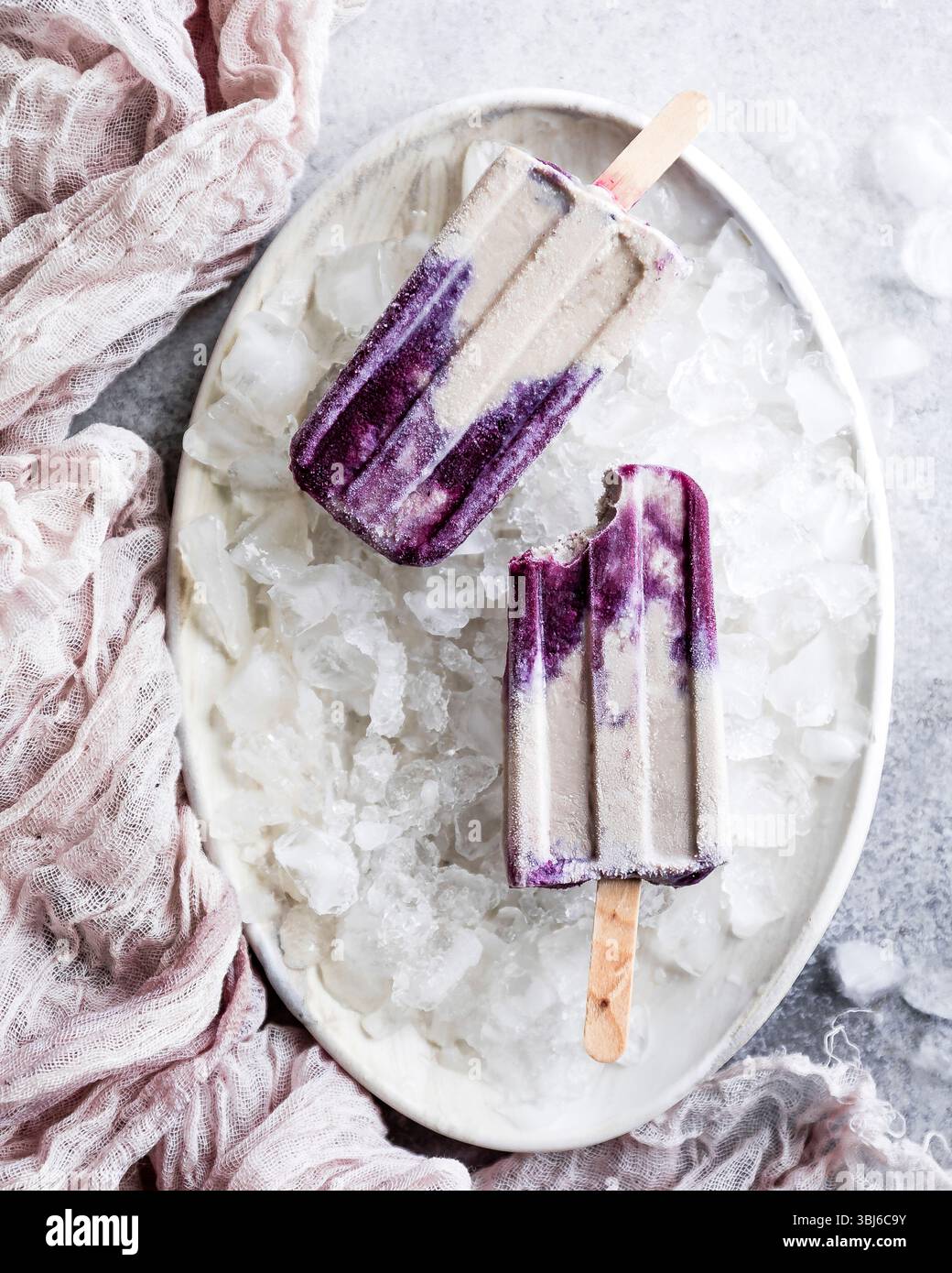 Deux popsicles violettes et crème sur un lit de glace dans un plat blanc, avec un chiffon rose à proximité. Banque D'Images