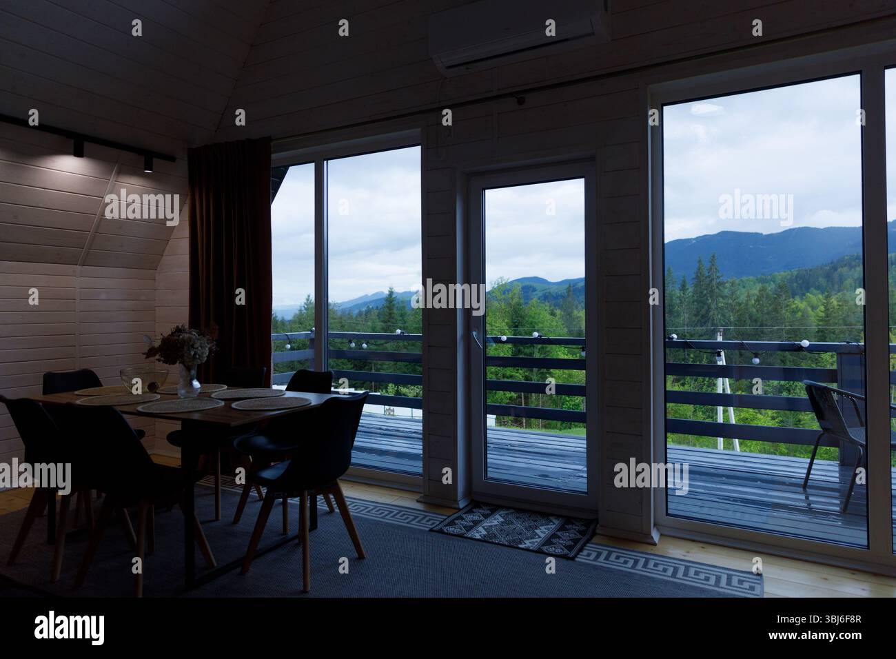 Un intérieur de cabine en bois invitant mettant en vedette une salle à manger avec des chaises élégantes, de grandes fenêtres panoramiques révélant des montagnes verdoyantes et un cadre tranquille Banque D'Images