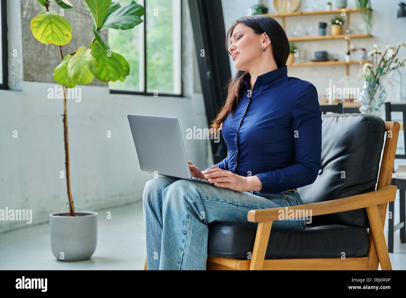 Femme fatiguée travaillant avec un ordinateur portable, ayant mal au cou, assise dans une chaise avec un ordinateur Banque D'Images