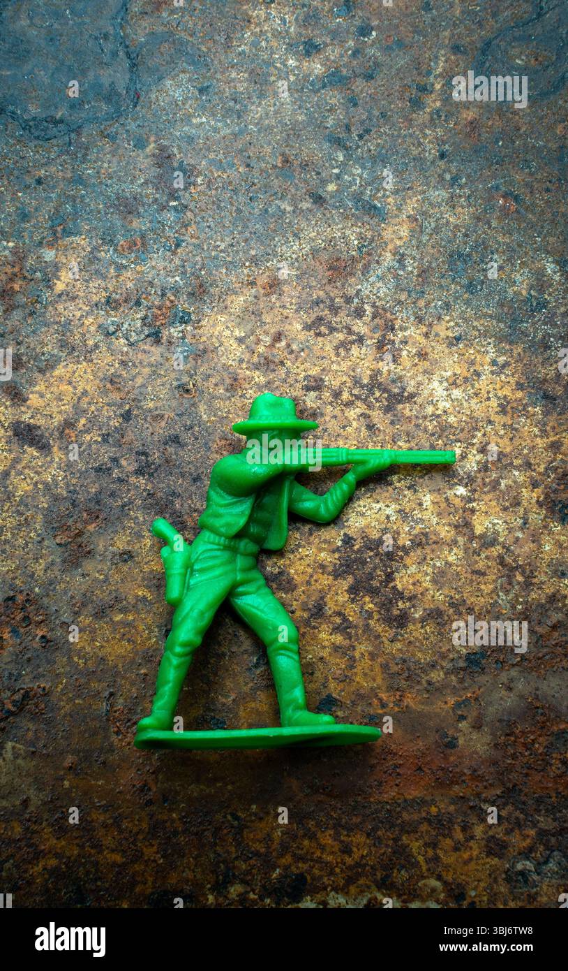 Un petit cow-boy jouet en plastique vert se tient fermement sur un fond texturé bleu et brun rustique. La figurine est équipée d'une arme Banque D'Images