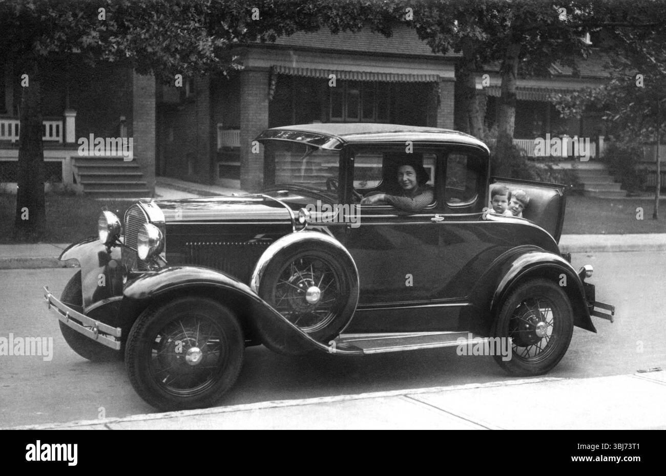 Femme dans les enfants de voiture vintage, années 1930, Une voiture vintage avec des passagers conduit le long d'une rue avec des maisons classiques, photo historique Banque D'Images
