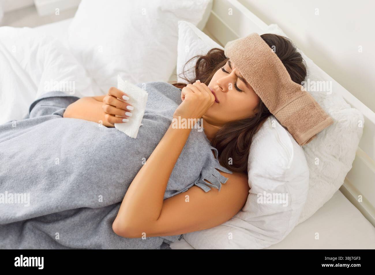Jeune femme malade avec une serviette mouillée sur la tête couchée dans le lit, souffrant du virus du rhume ou de la grippe et de la toux. Banque D'Images