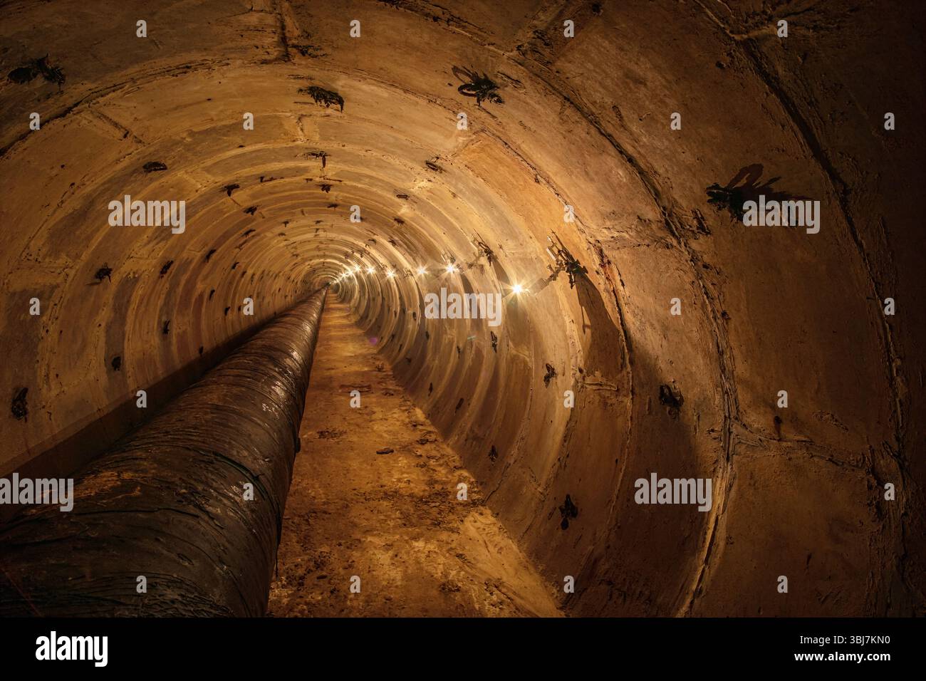 Long tunnel rond souterrain en béton avec un tuyau, éclairé par des lampes à incandescence. Tunnel souterrain d'une conduite de chauffage ou d'un système d'alimentation en eau. Banque D'Images