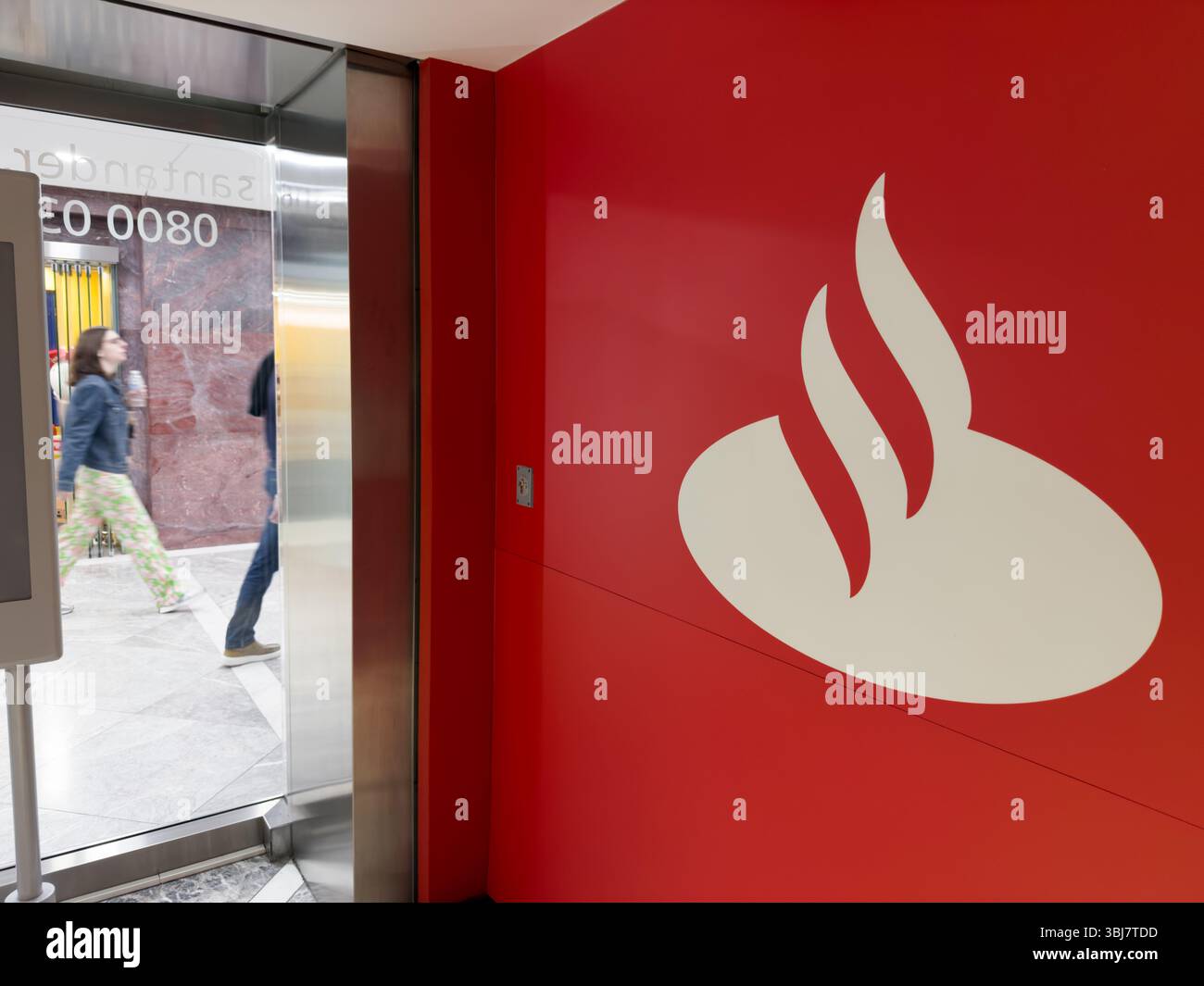 Le client entre dans la succursale de Santander avec le logo emblématique de la flamme blanche sur le mur rouge audacieux. La présence frappante de la marque accueille les visiteurs à l'entrée. Banque D'Images