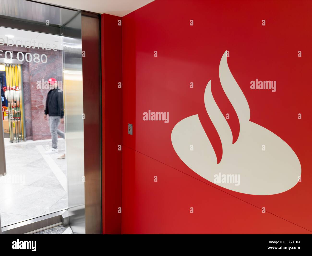 Le client entre dans la succursale de Santander avec le logo emblématique de la flamme blanche sur le mur rouge audacieux. La présence frappante de la marque accueille les visiteurs à l'entrée. Banque D'Images