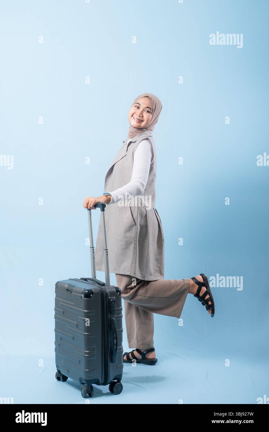 Une femme tient une valise et sourit. La valise est argentée et a des roues. La femme porte une chemise blanche et une écharpe. Concept de voyage et Banque D'Images