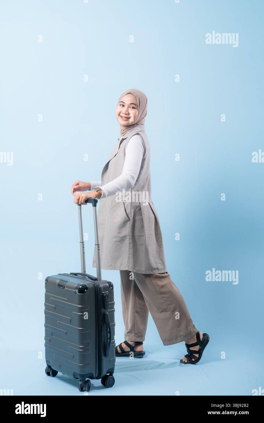 Une femme tient une valise et sourit. La valise est argentée et a des roues. La femme porte une chemise blanche et une écharpe. Concept de voyage et Banque D'Images