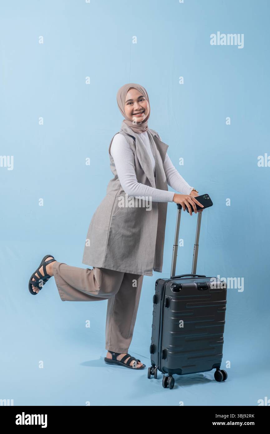 Une femme portant un gilet gris et un pantalon bronzé sourit et tient une valise. La valise est noire et a des roues Banque D'Images