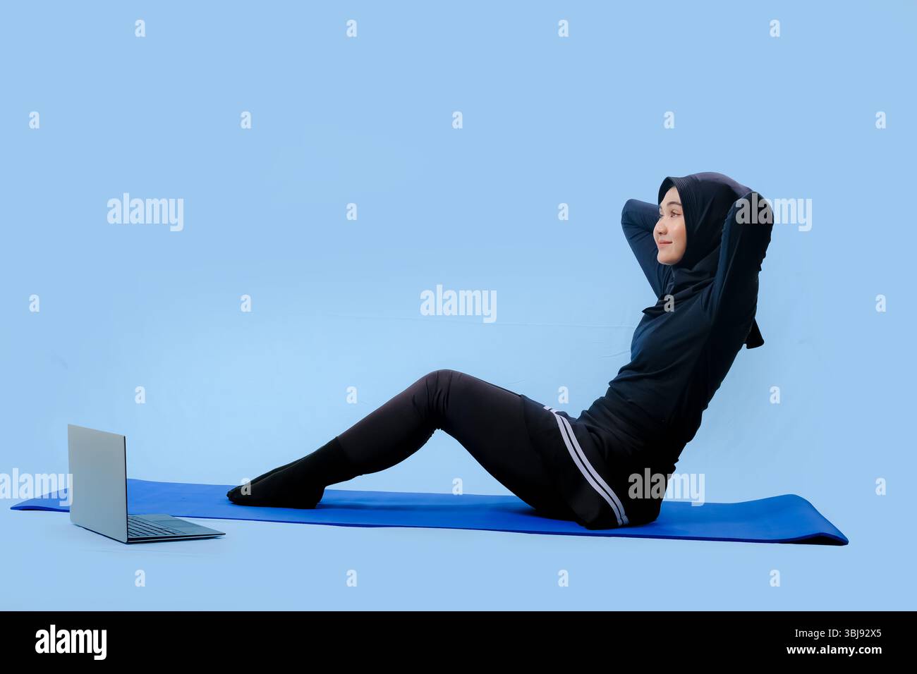 Une femme est assise sur un tapis de yoga avec un ordinateur portable sur ses genoux. Elle porte un top noir et un pantalon noir Banque D'Images