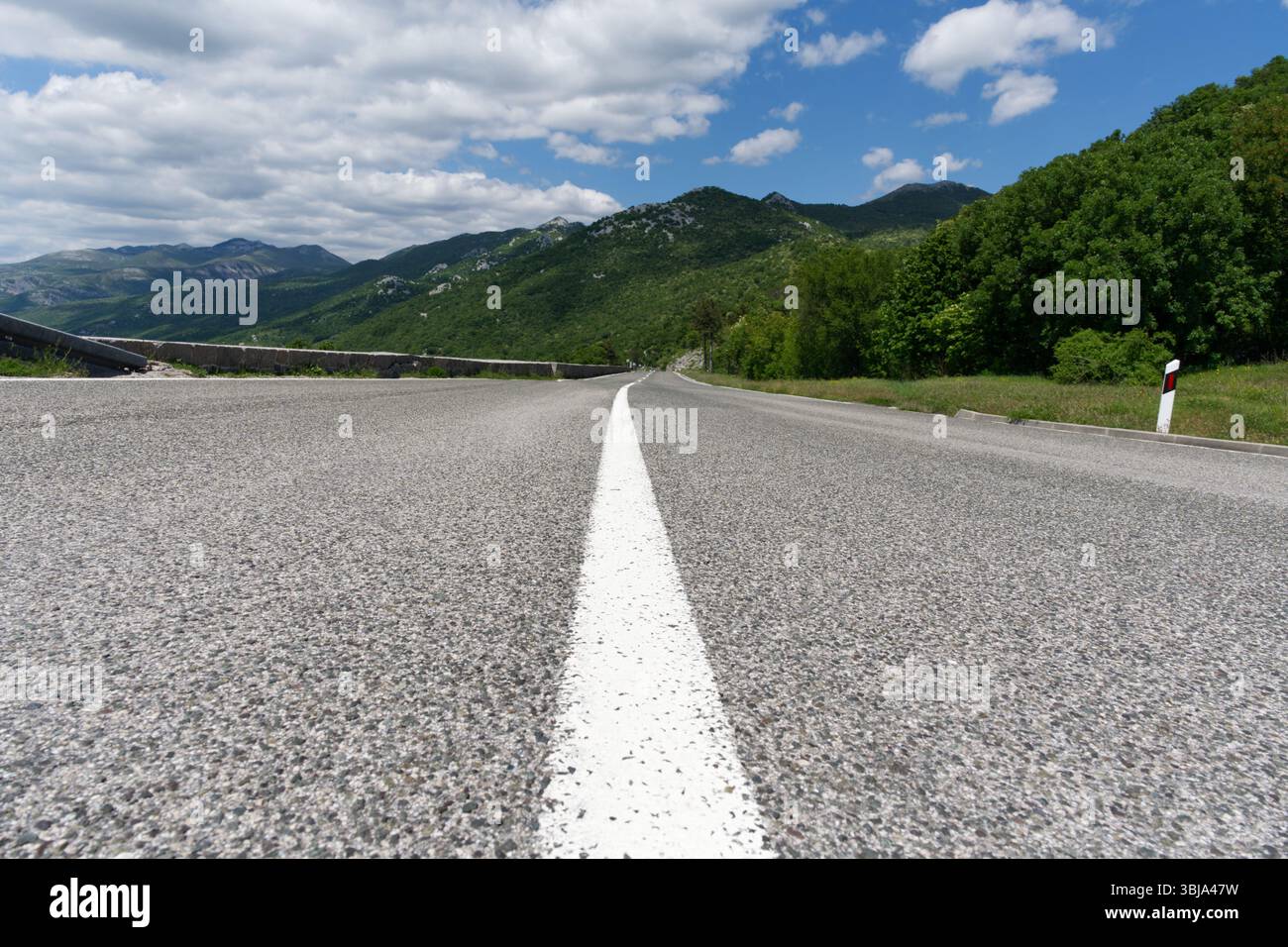Une vue en bas angle capture une route pavée s'étendant vers des montagnes vertes lointaines sous un ciel partiellement nuageux, avec une ligne centrale blanche guidant l'œil. Banque D'Images
