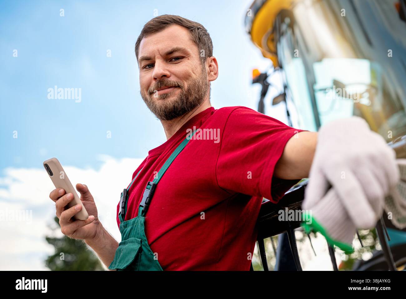 Portrait d'un ouvrier agricole souriant confiant avec smartphone à côté de la moissonneuse-batteuse Banque D'Images