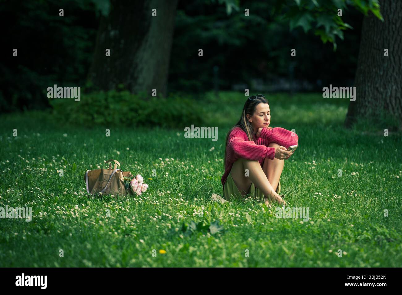 Jeune fille assise dans l'herbe seule et regardant le téléphone dans un parc public en été Banque D'Images