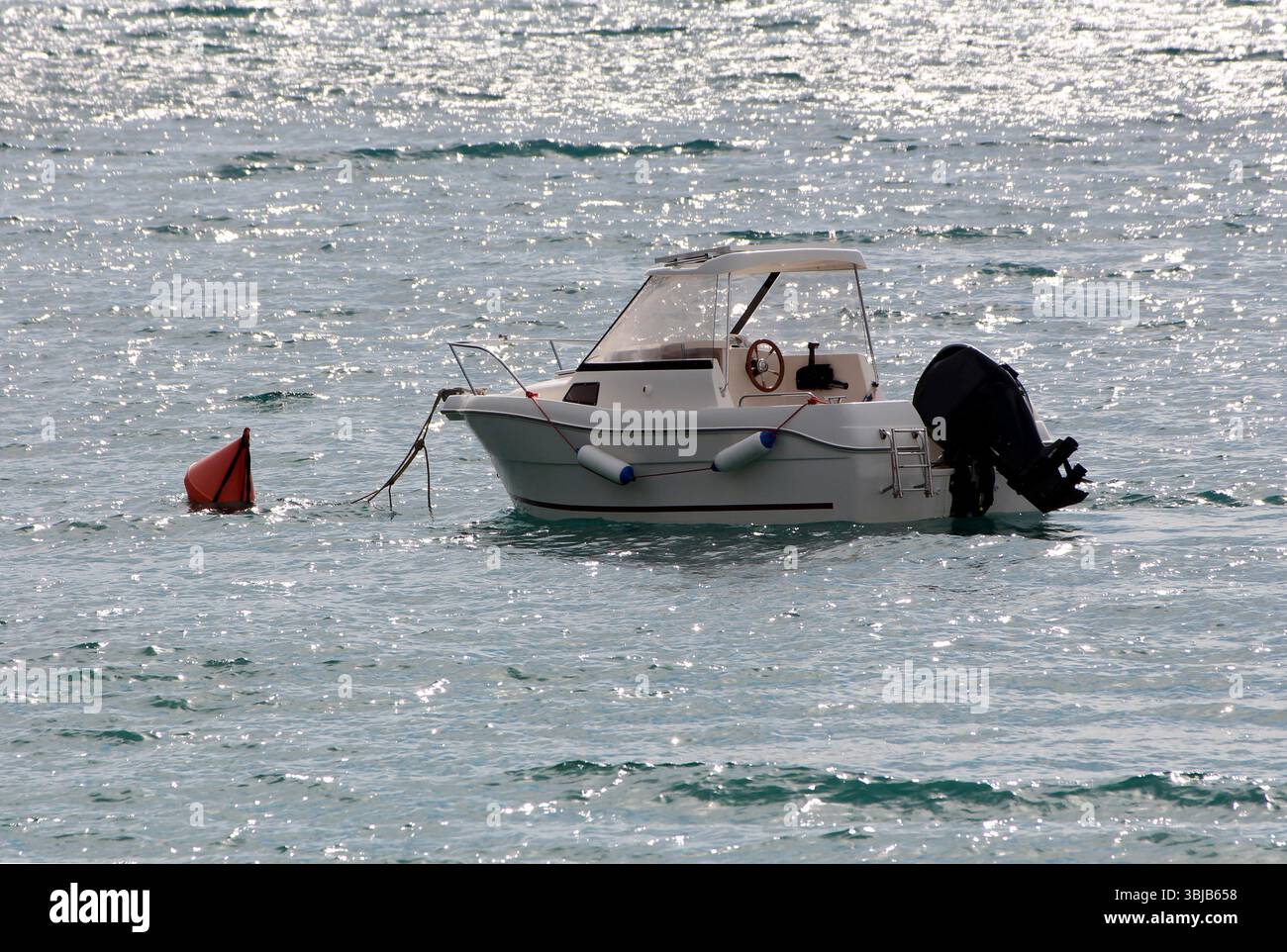 Bateau à moteur blanc avec cabine et moteur hors-bord flotte sur l'eau pétillante, attaché à une bouée rouge, avec des vagues douces et des reflets de la lumière du soleil créant Banque D'Images