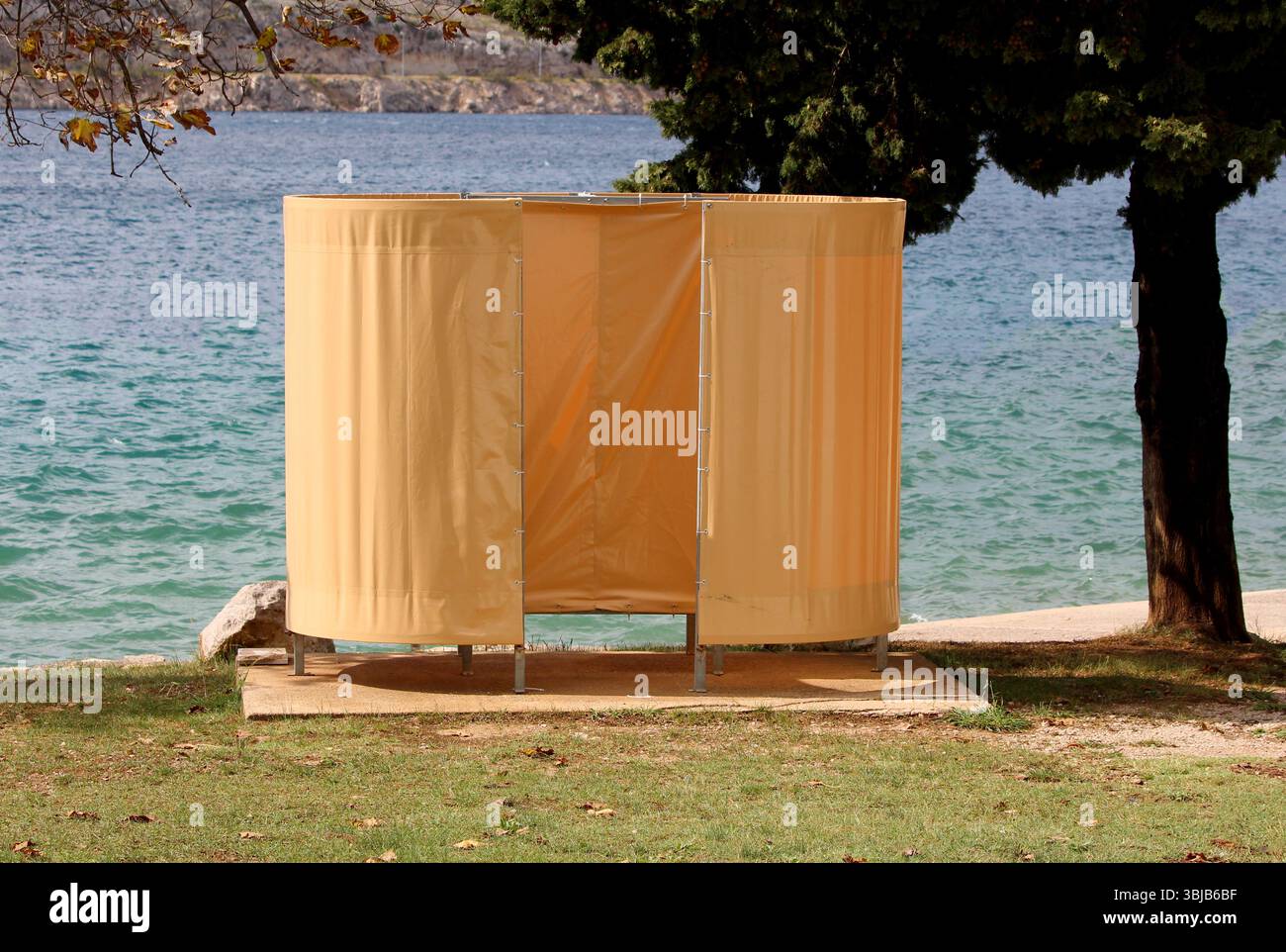 Cabine à langer jaune portable de plage avec panneaux incurvés se dresse sur une dalle de béton au bord de l'eau, ombragée par des arbres, offrant l'intimité pour les nageurs Banque D'Images