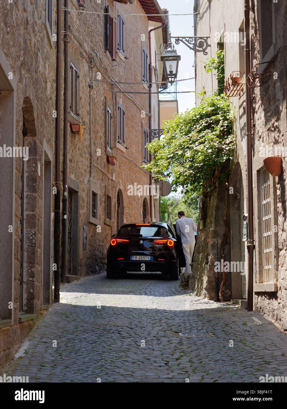Homme habillé intelligemment monte dans une voiture Alfa Romeo avec des lumières allumées dans une rue pittoresque étroite à Montefiascone, région du Latium, Italie. 15 juin 2025 Banque D'Images