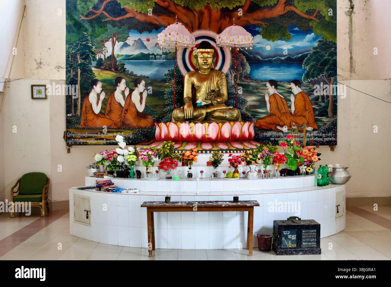 Autel du temple bouddhiste Srimangal à Kuakata, Bangladesh, avec un Bouddha doré devant une murale, des offrandes florales et une boîte de dons. Banque D'Images