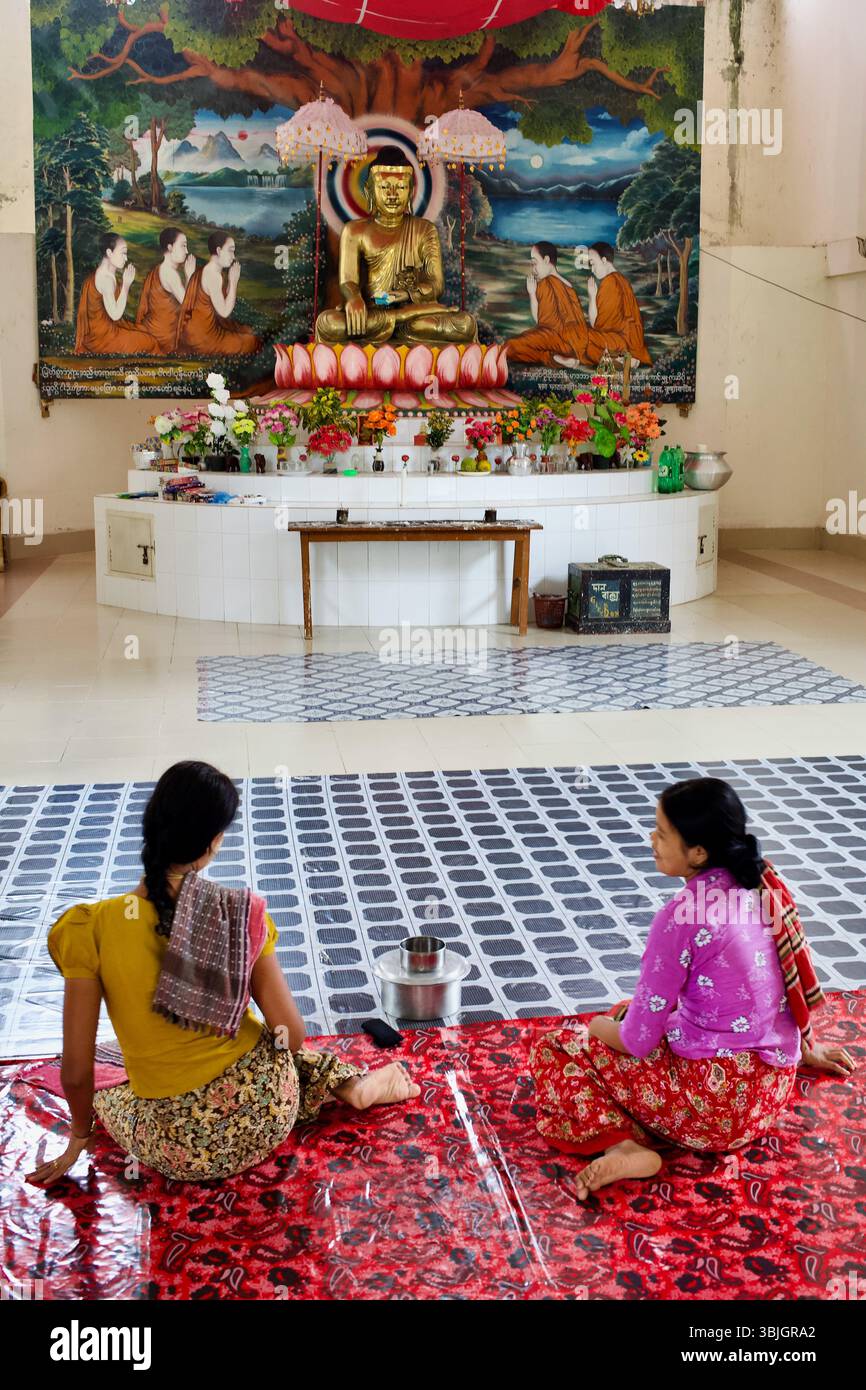 Des femmes assises en prière au temple bouddhiste Srimangal à Kuakata, Bangladesh, face à une murale vive et à Bouddha doré assis. Banque D'Images