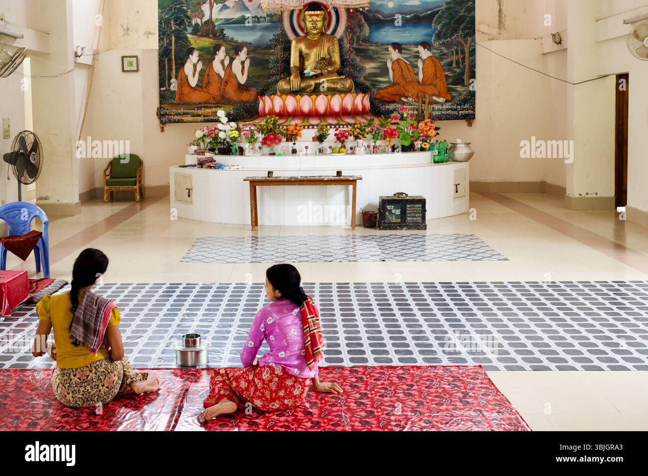 Des femmes assises en prière au temple bouddhiste Srimangal à Kuakata, Bangladesh, face à une murale vive et à Bouddha doré assis. Banque D'Images
