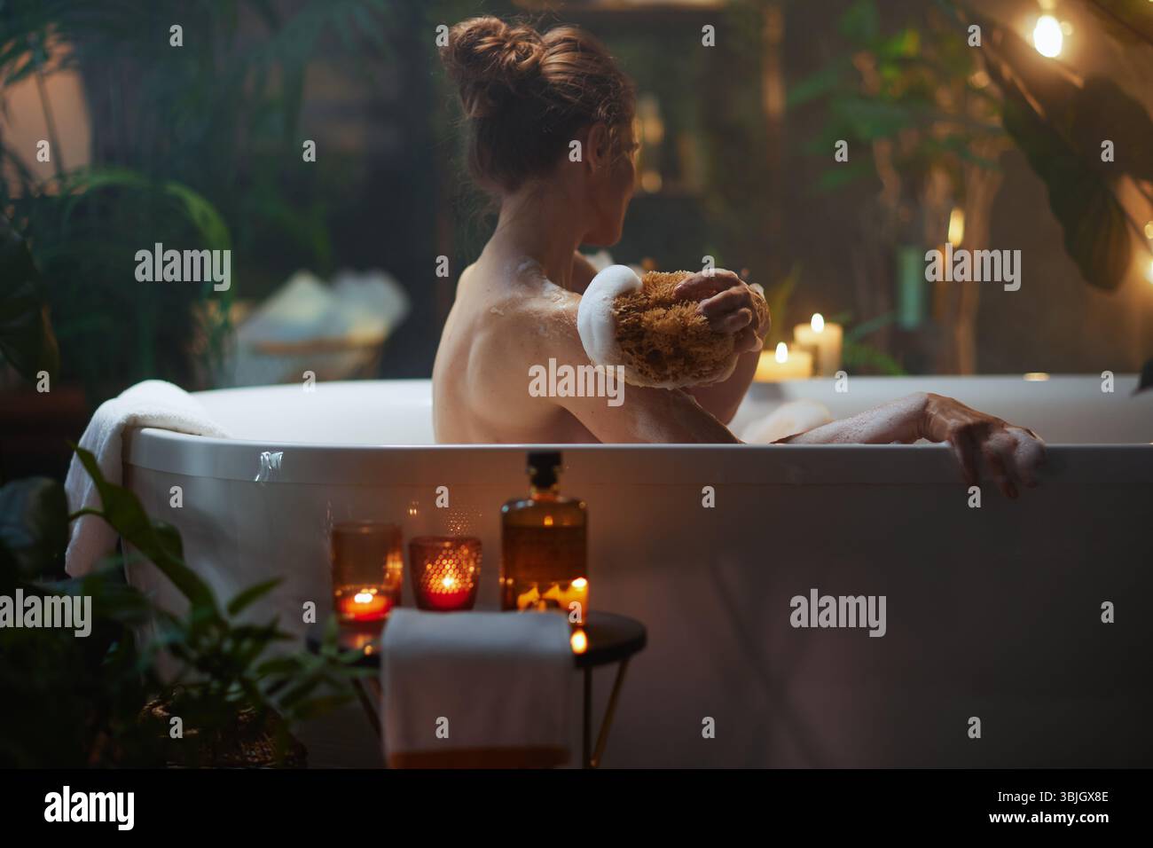 Une femme à la peau claire se livre à un luxueux rituel d'auto-soin, frottant doucement son épaule avec une éponge naturelle dans une baignoire chaude et éclairée aux chandelles Banque D'Images