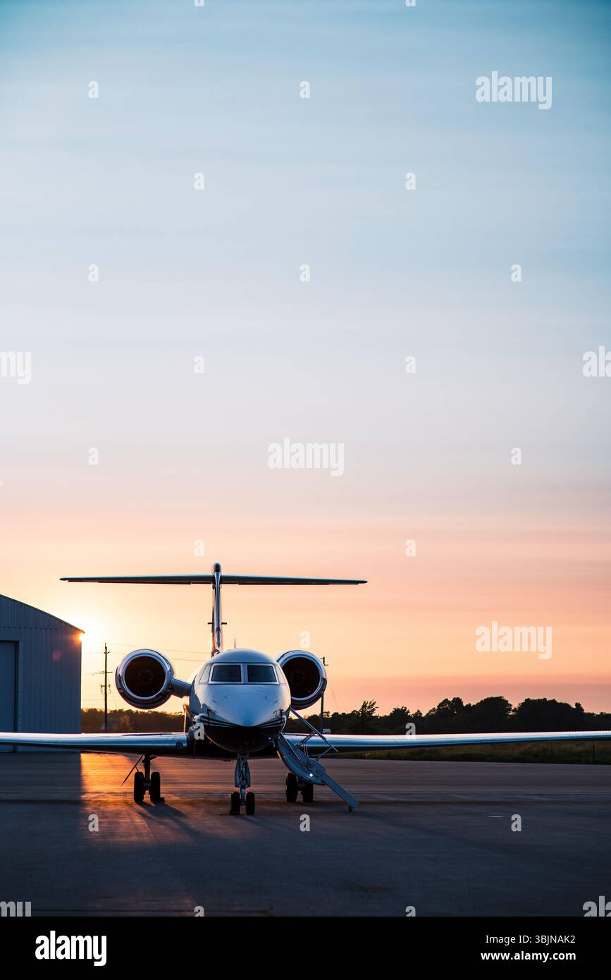 Jet privé silhouetté contre un coucher de soleil doré, garé sur le tarmac à côté d'un hangar. Départs en soirée et voyages de luxe. Banque D'Images
